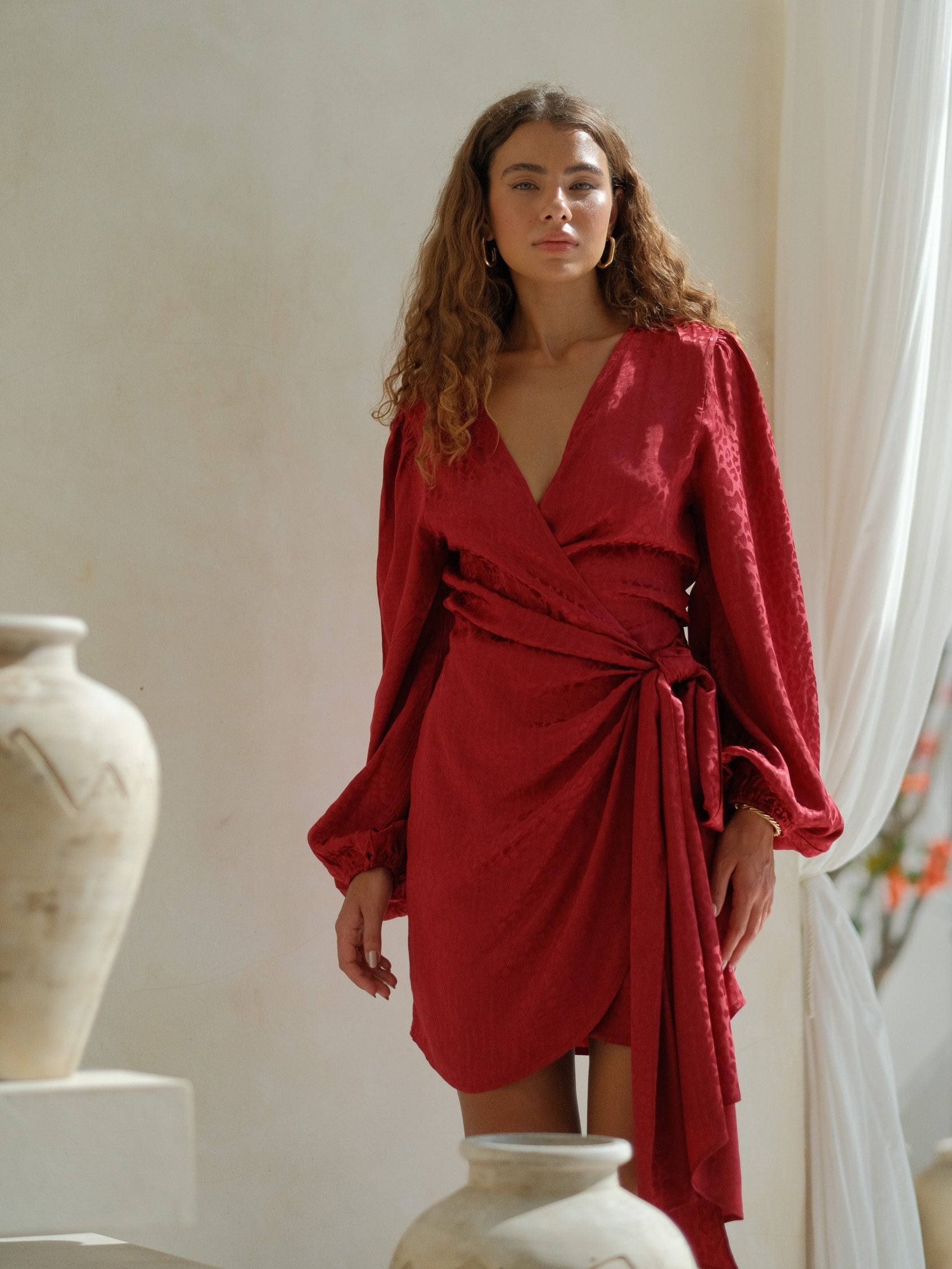 Burgundy jacquard wrap mini dress featuring a wrap-style neckline, long voluminous sleeves, and a side tie detail with a softly draped silhouette. Lightweight textured fabric with a subtle sheen, perfect for statement evening and elevated occasion wear.