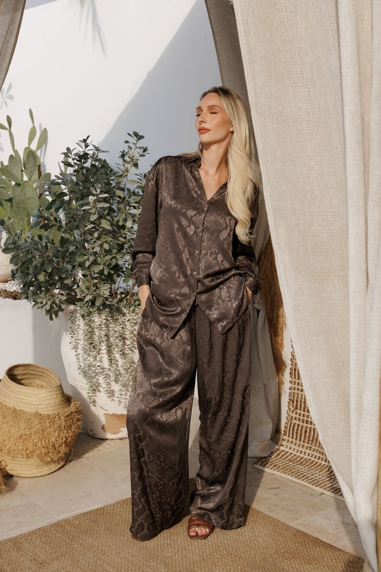 Elegant brown printed satin resortwear set with oversized shirt and relaxed trousers, styled against a minimal wall with soft tropical shadows.