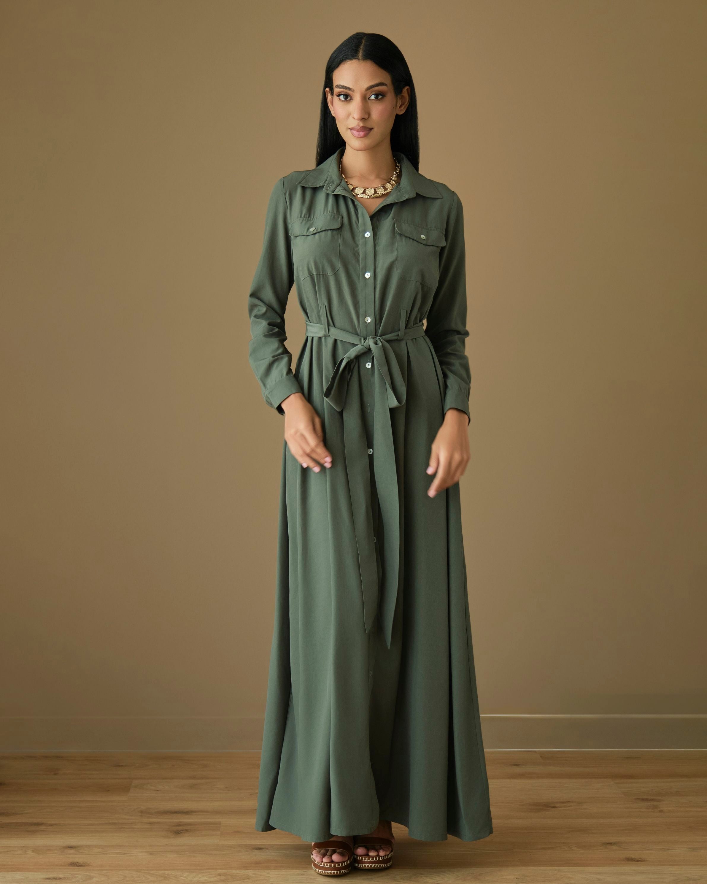 Model wearing a sage green maxi shirt dress with a collared neckline, button-front closure, long sleeves, flap chest pockets, and a matching self-tie waist belt.