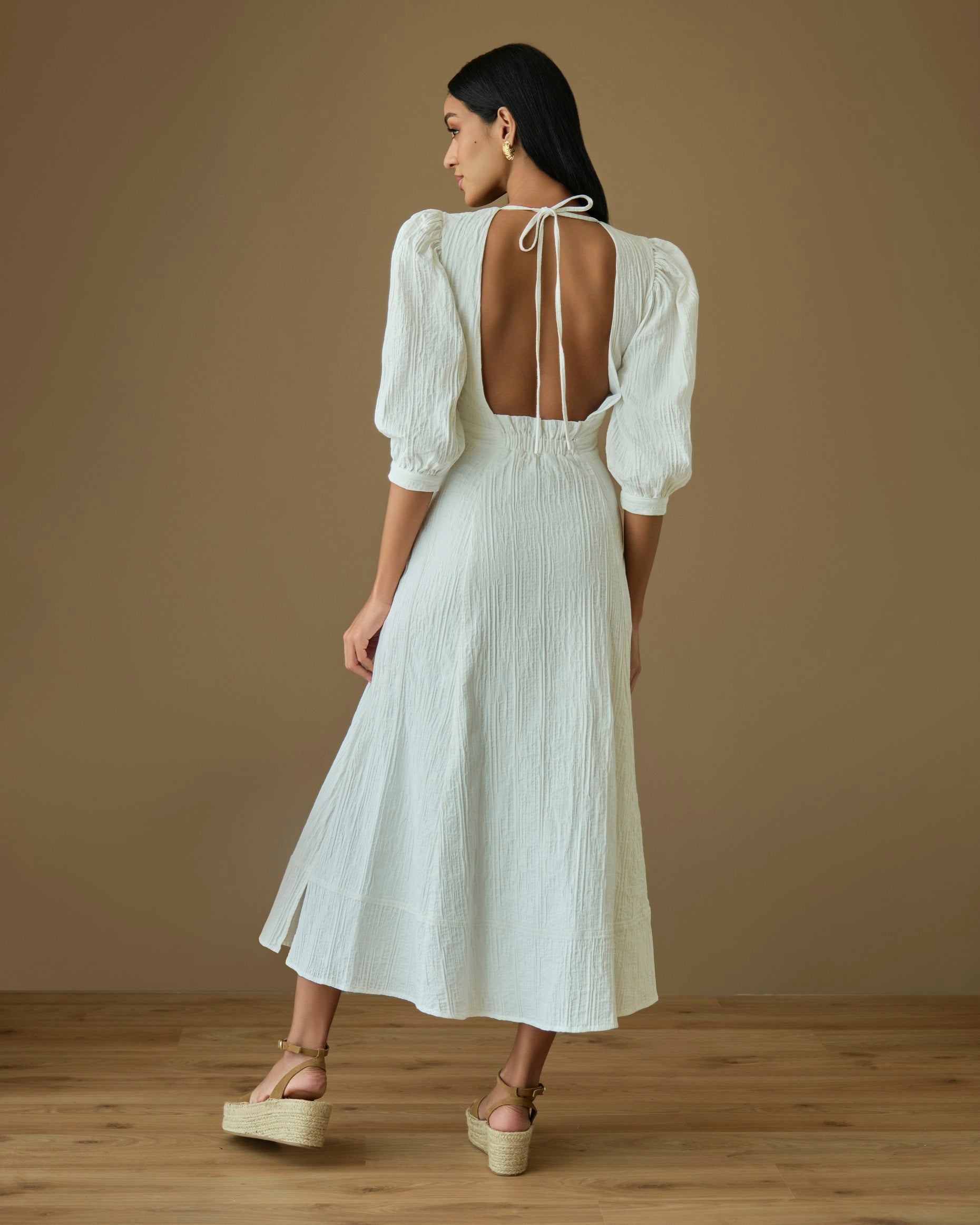 Model wearing the Nalla cotton dress, featuring an open back and midi length, styled on a wooden floor.