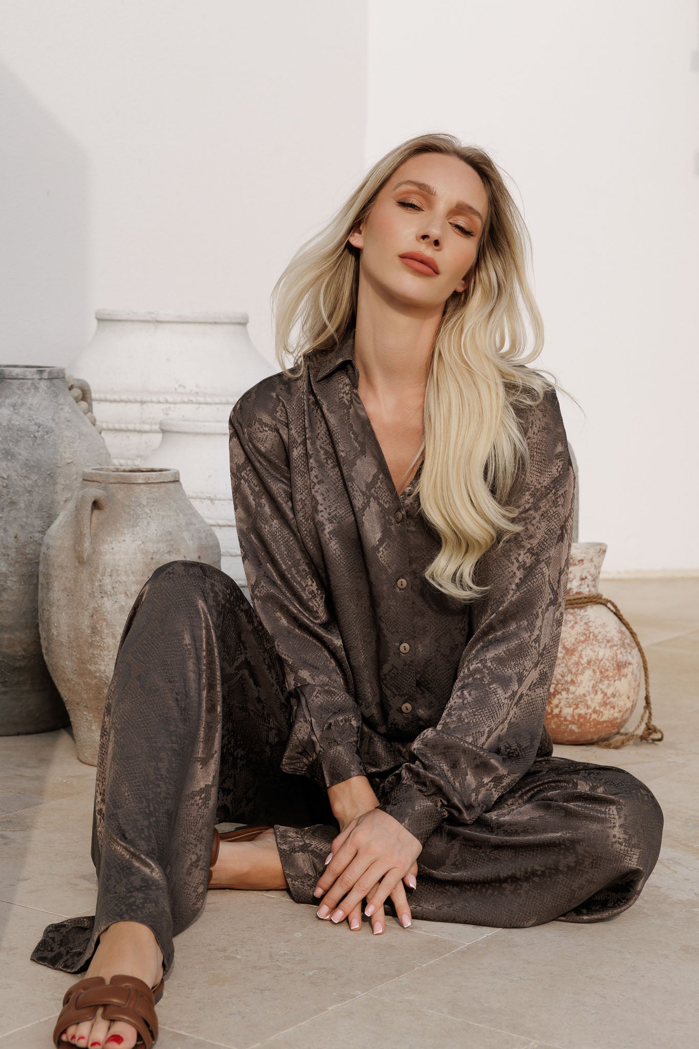Elegant brown printed satin resortwear set with oversized shirt and relaxed trousers, styled against a minimal wall with soft tropical shadows.