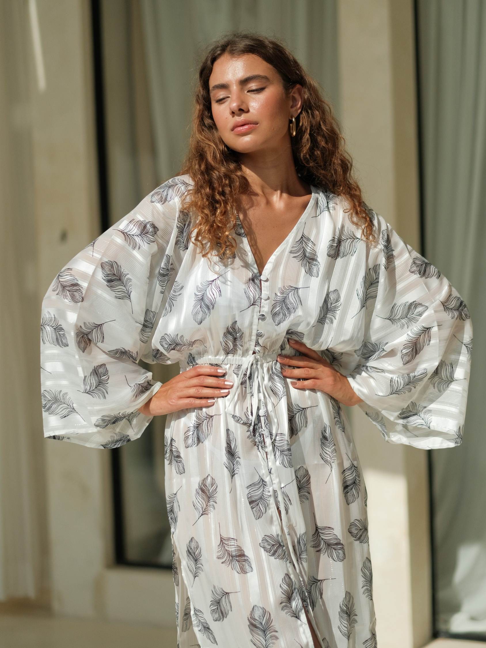 White chiffon cover-up featuring a soft feather print, long sleeves, and a relaxed flowing silhouette with a front button detail and high side slit. Lightweight, sheer chiffon fabric with an airy drape, perfect for effortless beach and resort layering.