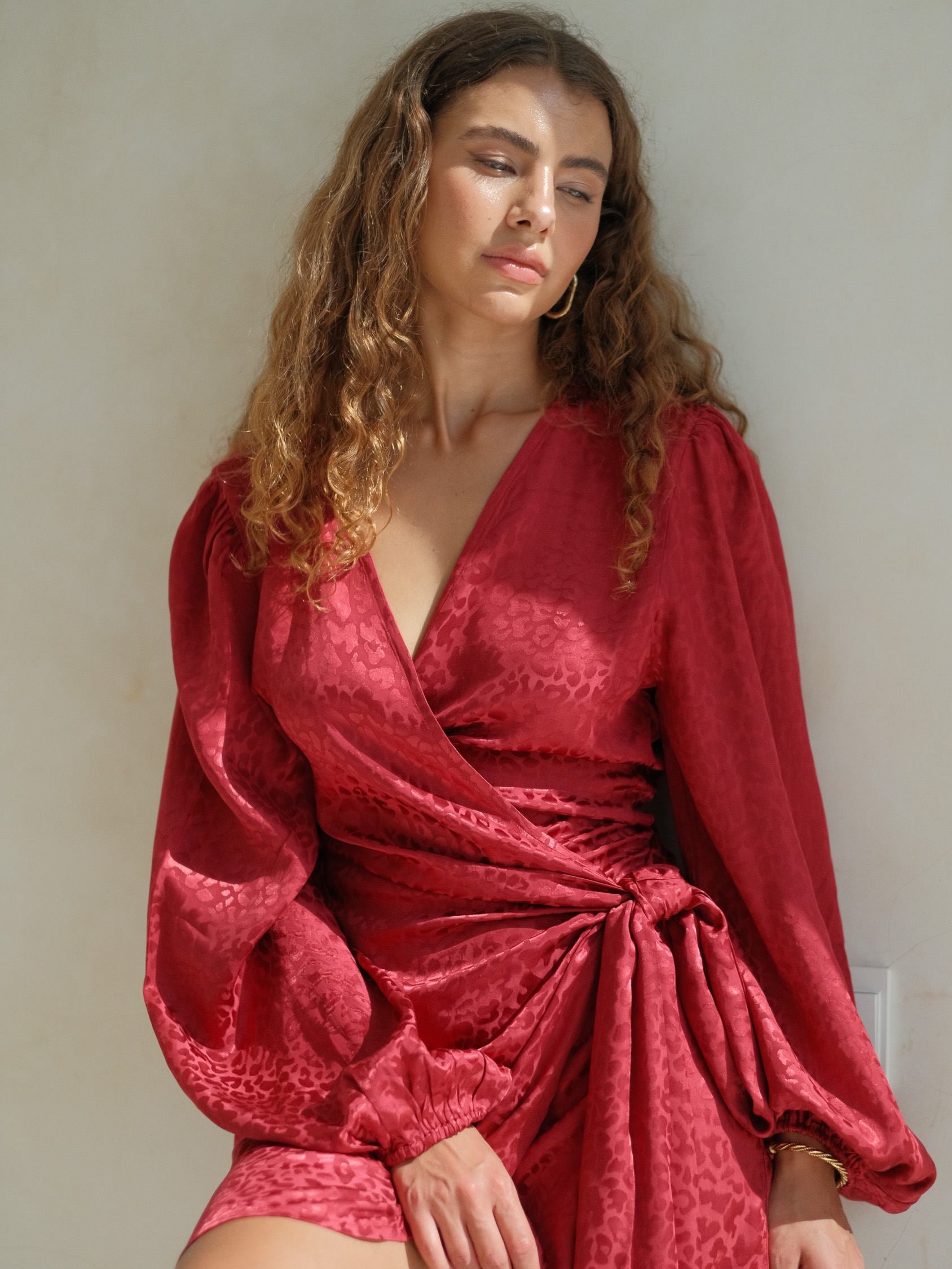 Burgundy jacquard wrap mini dress featuring a wrap-style neckline, long voluminous sleeves, and a side tie detail with a softly draped silhouette. Lightweight textured fabric with a subtle sheen, perfect for statement evening and elevated occasion wear.