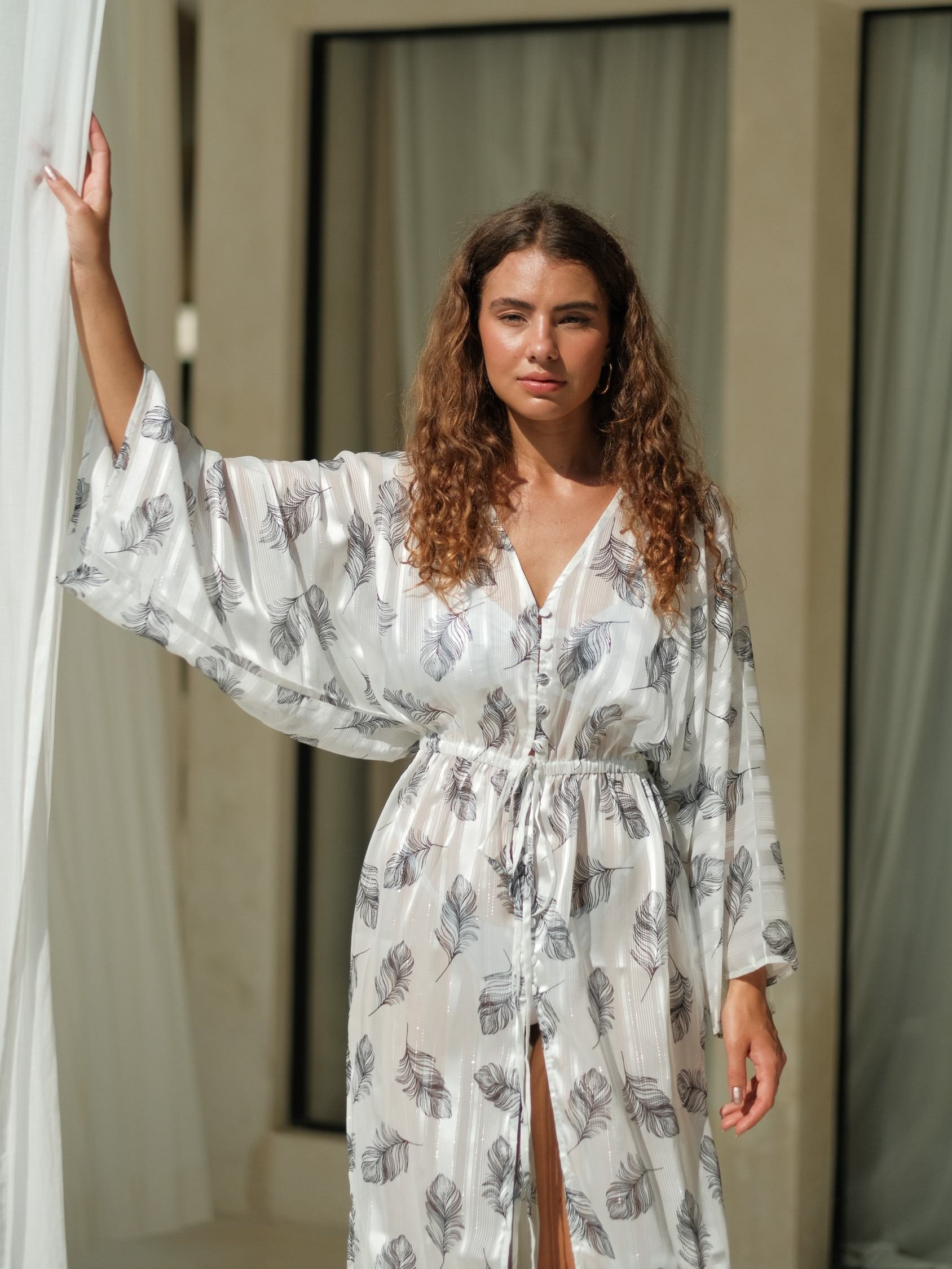 White chiffon cover-up featuring a soft feather print, long sleeves, and a relaxed flowing silhouette with a front button detail and high side slit. Lightweight, sheer chiffon fabric with an airy drape, perfect for effortless beach and resort layering.